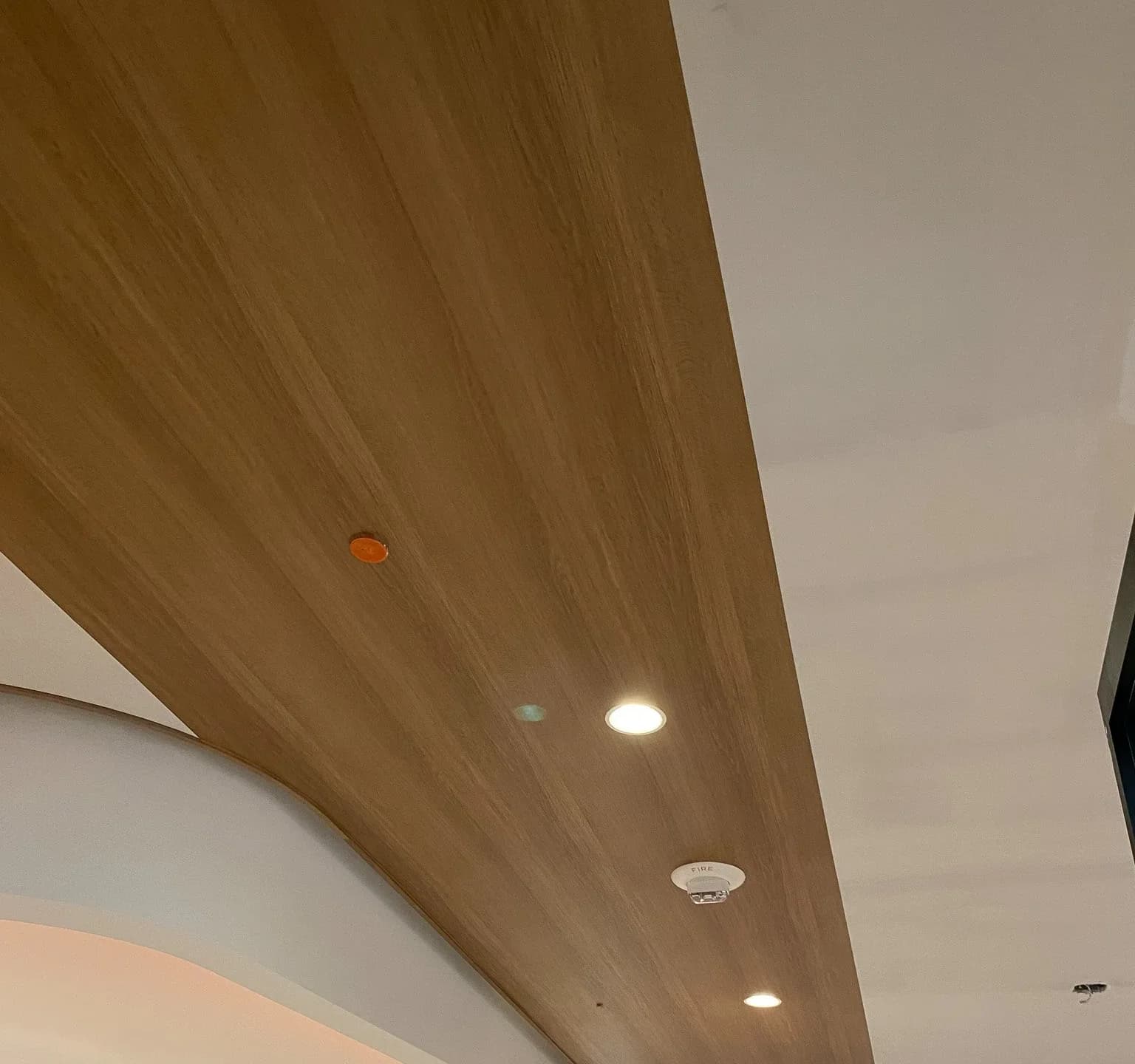 Wood-grain architectural film wrapping a curved ceiling soffit, cut in around downlights and a fire alarm, installed by Wrap Trap