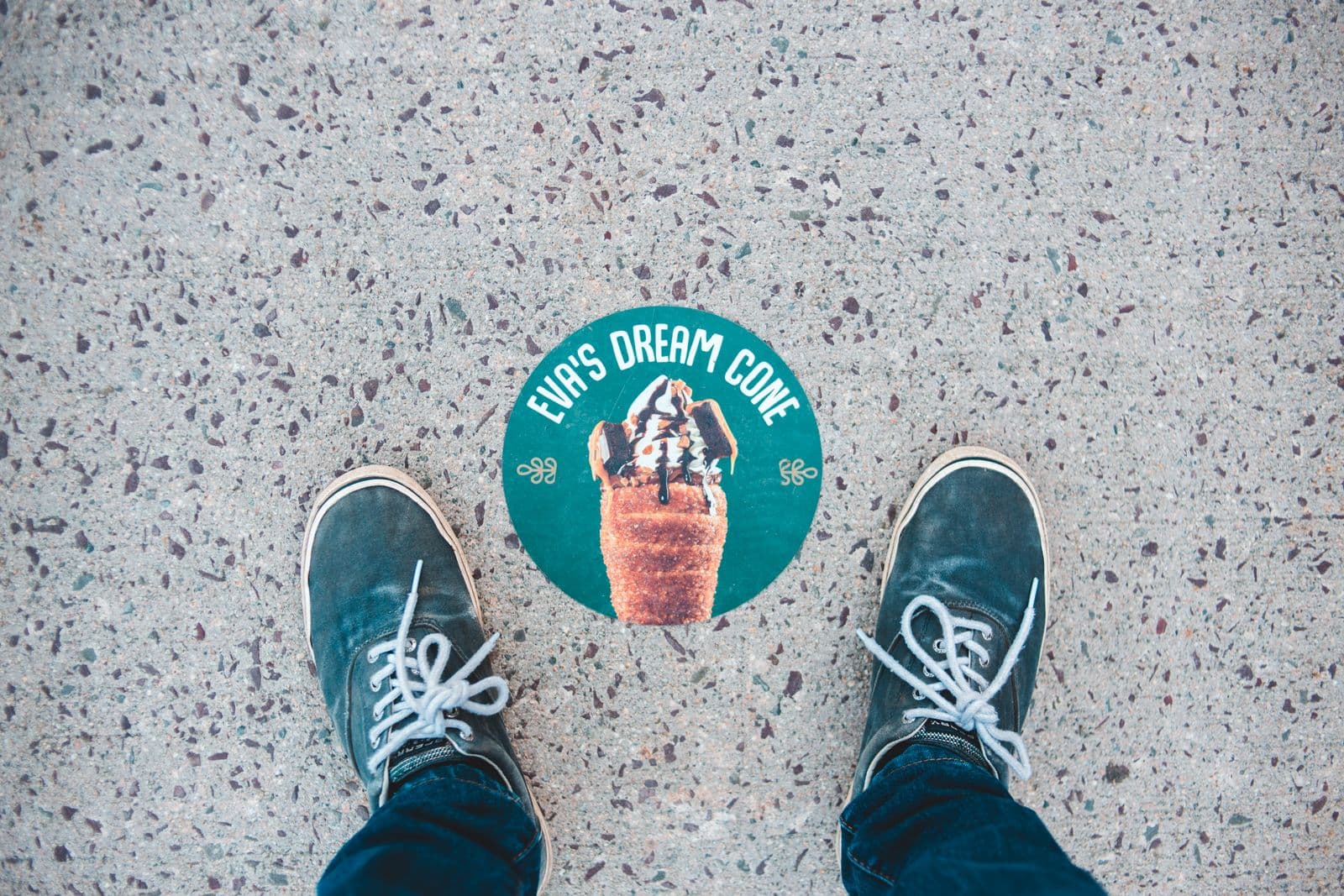 Round printed ice-cream-shop decal applied to a concrete sidewalk, seen from above with sneakers standing beside it