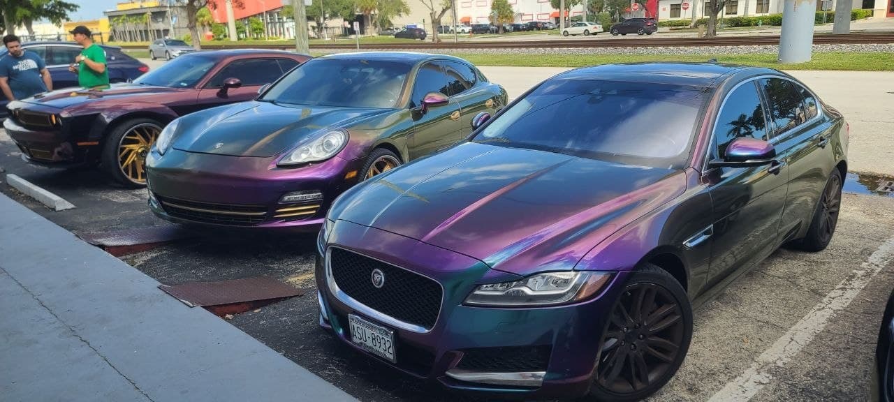Three color-shift wrapped cars in daylight — an iridescent purple-teal Jaguar XF in front, a purple-green Porsche Panamera and a dark color-shift muscle car behind it, each showing two colors in the same frame.